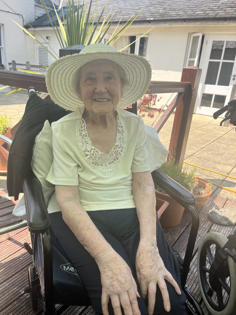 Elderly woman in a wheelchair smiling outdoors, wearing a sun hat.