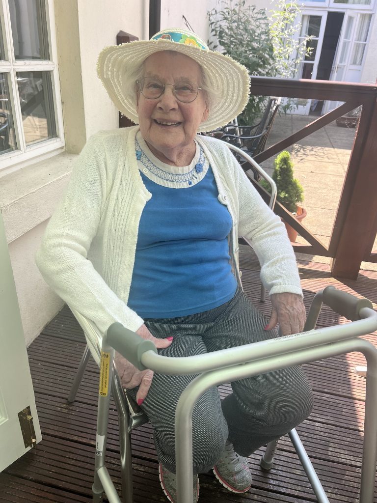 Elderly woman sitting with a walker, smiling and wearing a sunhat.