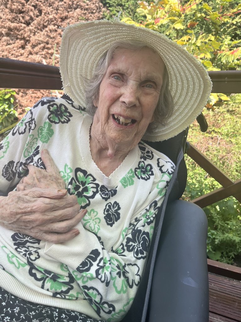 Elderly woman sitting with a walker, smiling and wearing a wide-brimmed hat.