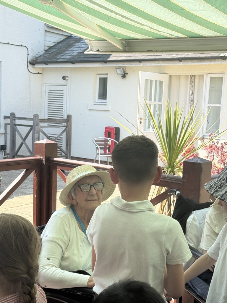 Elderly woman in a hat sitting outside, talking with children gathered around her.