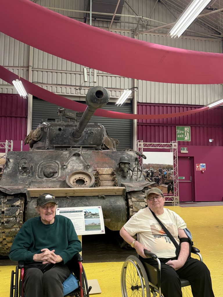 Two men in wheelchairs posing in front of a large historic tank inside a museum.