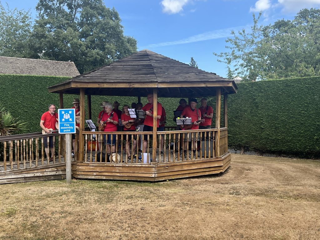 Group of musicians in red shirts playing ukuleles in a wooden gazebo outdoors.