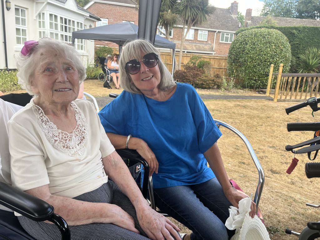 Two women smiling outdoors, one elderly seated in a wheelchair and the other beside her.