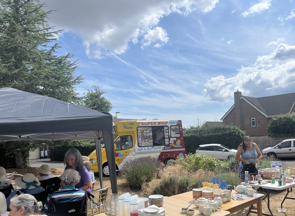 Residents sit under a gazebo at a summer fair with drinks table and ice cream van nearby.