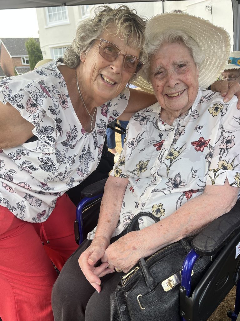 Two women smiling together, one seated in a wheelchair, sharing a warm moment outdoors.