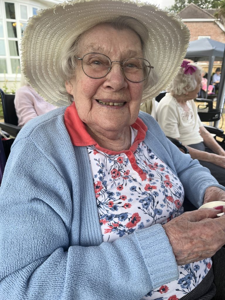 Close-up of an elderly woman in a sunhat and glasses smiling while seated outside.