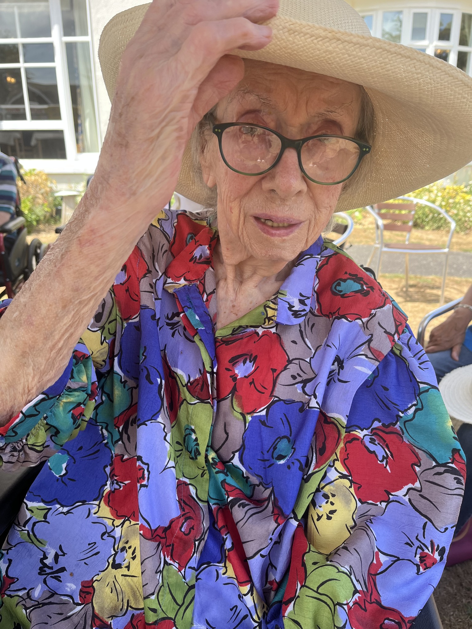 Elderly woman in a colorful shirt adjusting her hat while looking at the camera.
