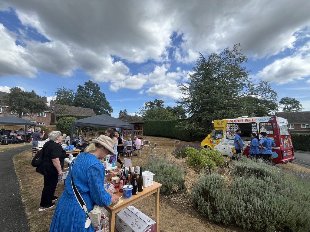 Outdoor summer fair with people browsing a table and an ice cream van nearby under a cloudy sky.