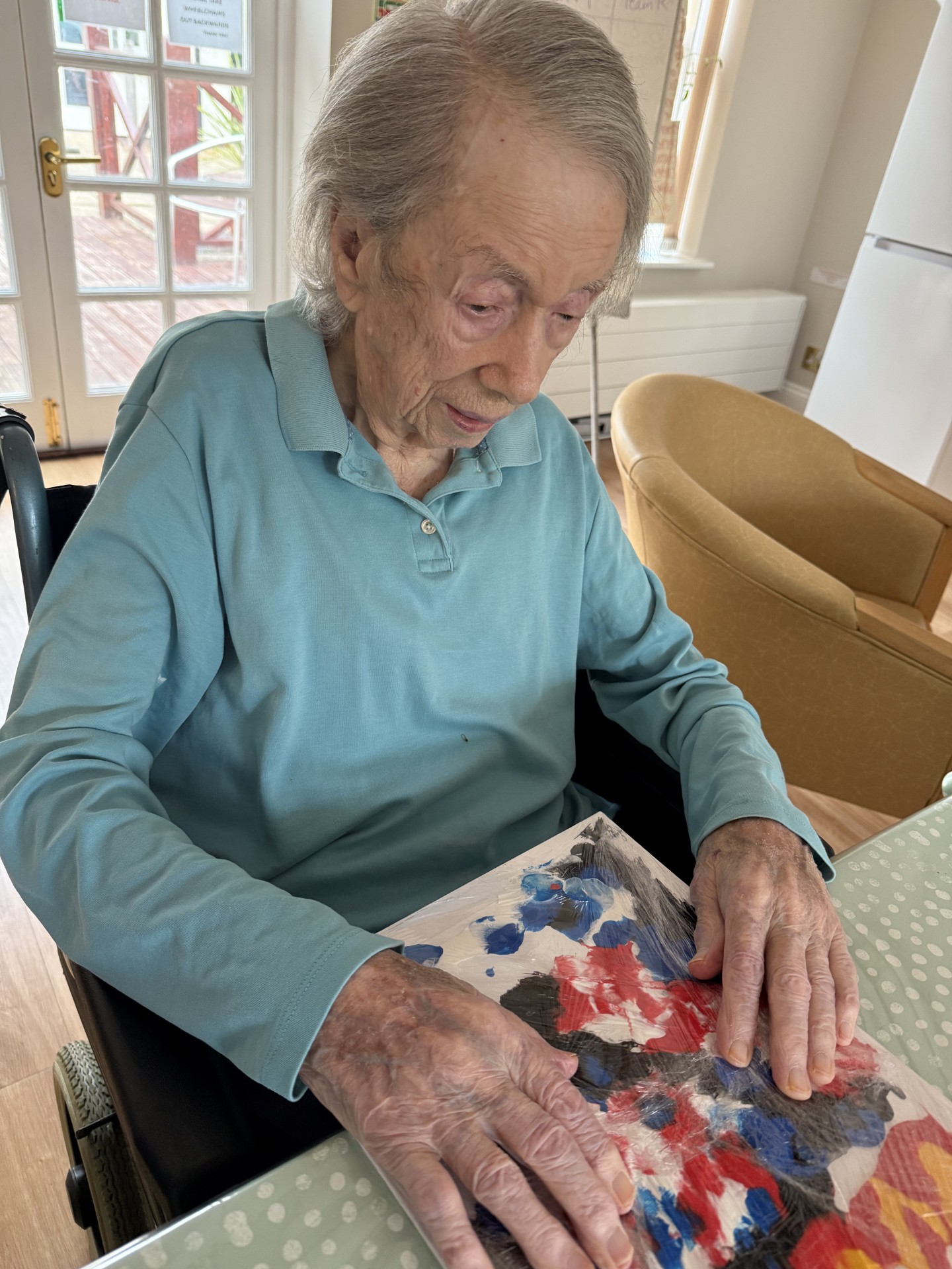 Elderly woman in a wheelchair creating colorful artwork on paper at a table.