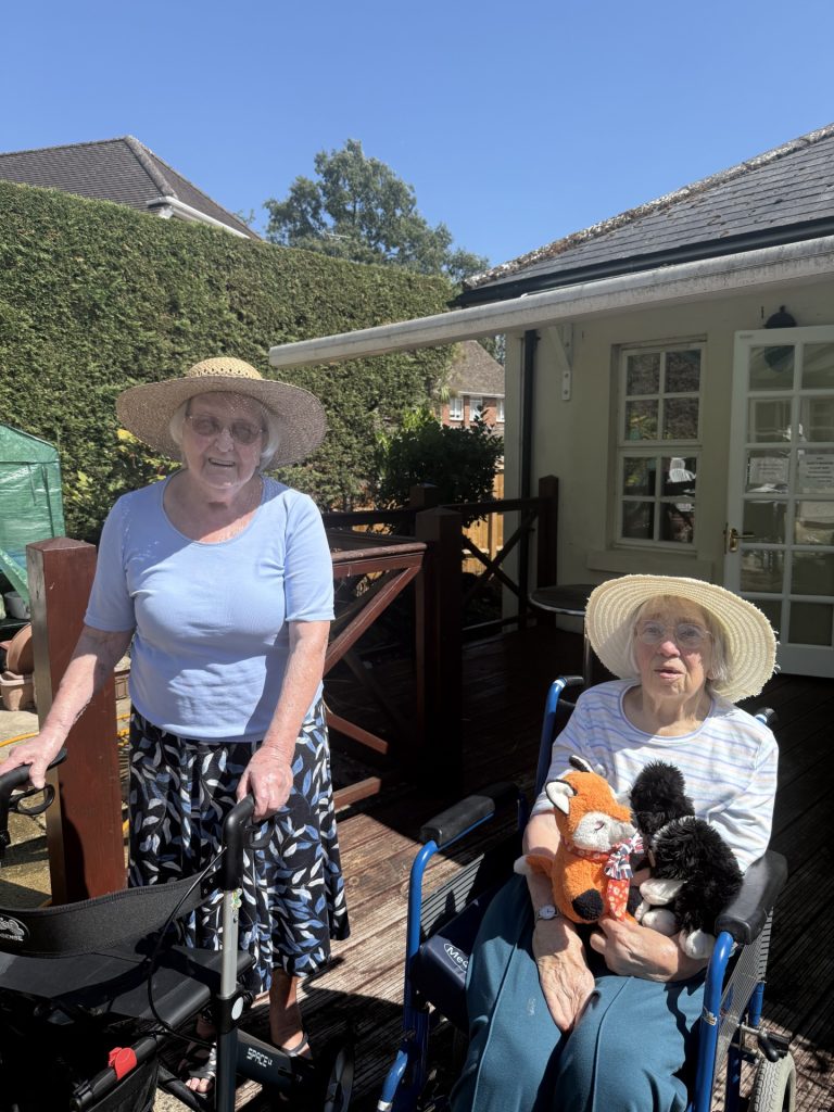 Elderly woman standing with a walker beside another woman in a wheelchair holding soft toys, both wearing sun hats outdoors.