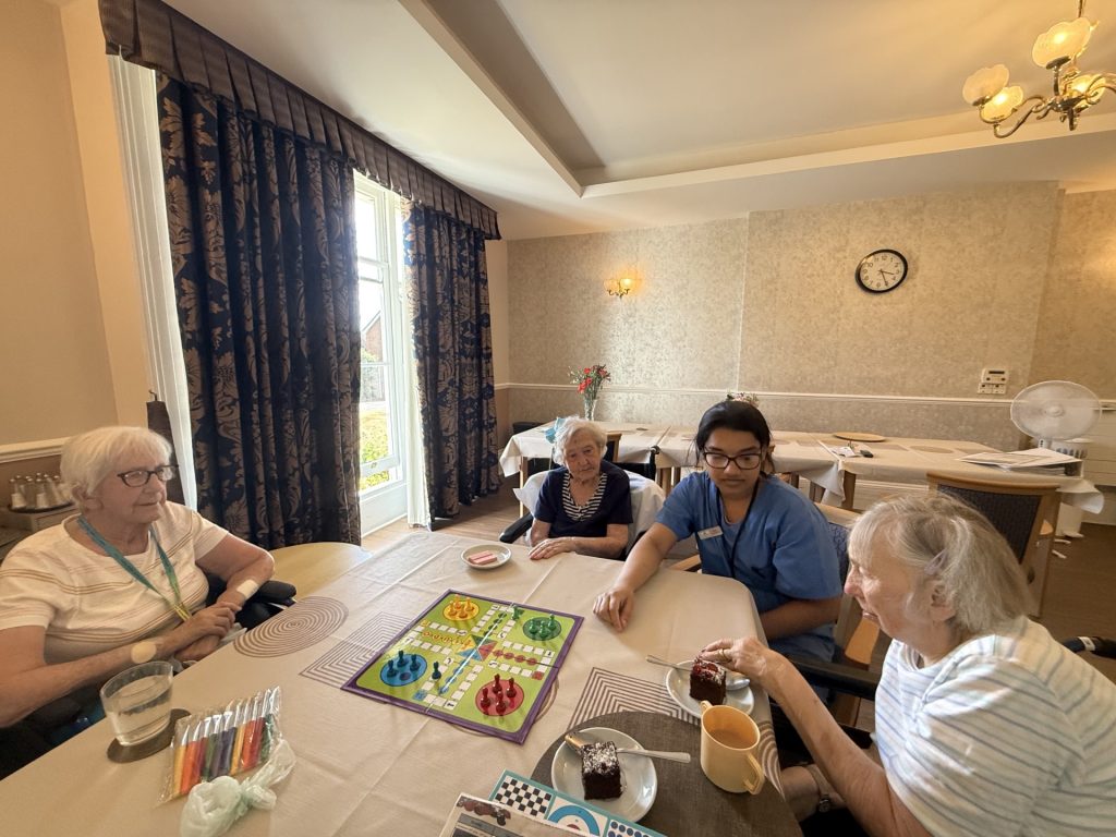 Group of elderly women and a caregiver playing a board game at a table with cake.