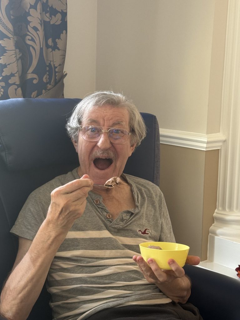 Elderly man smiling while eating dessert from a spoon.