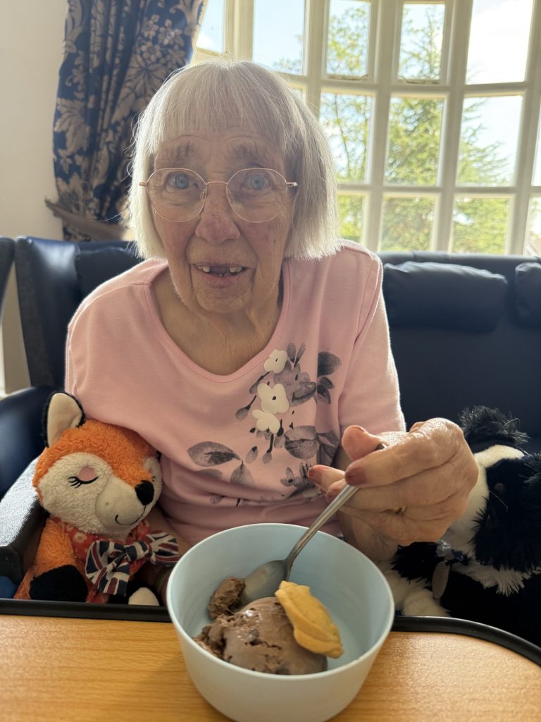 Elderly woman with glasses holding a bowl of ice cream, with soft toys beside her.