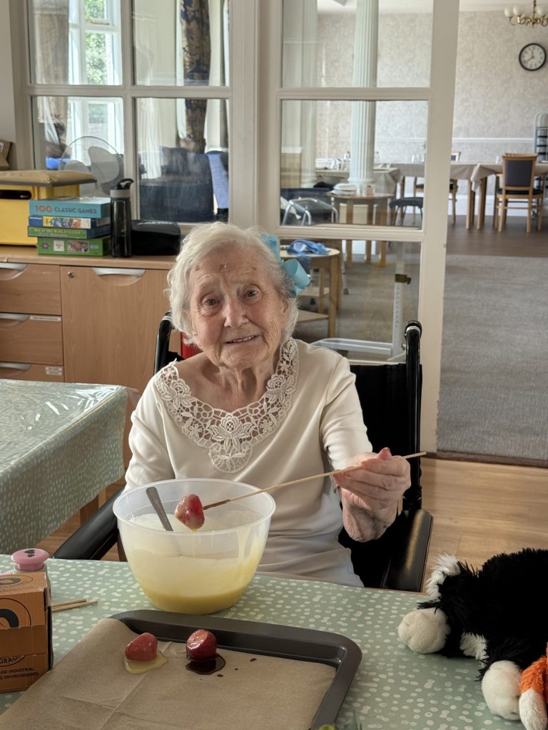 Elderly woman in a wheelchair dipping a strawberry into a bowl of melted chocolate.