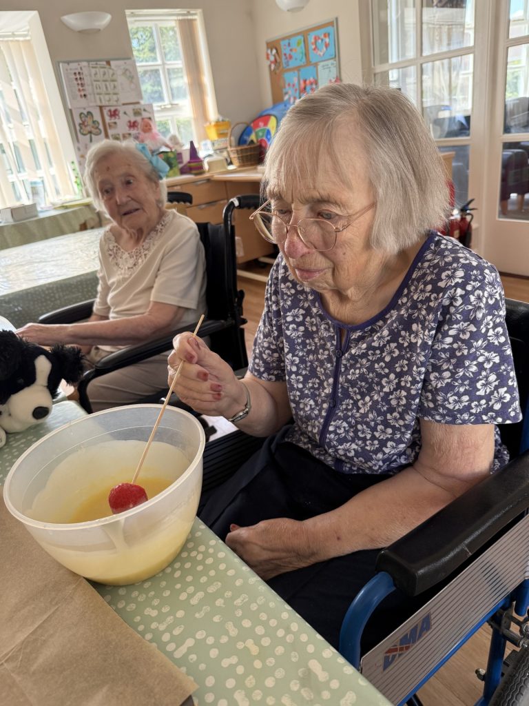 Two elderly women in wheelchairs dipping strawberries into chocolate at a table.