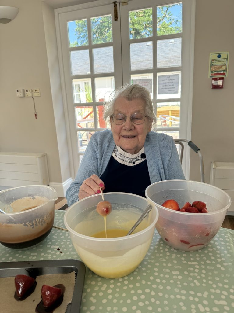 Elderly woman smiling while dipping a strawberry into chocolate, with bowls of fruit nearby.