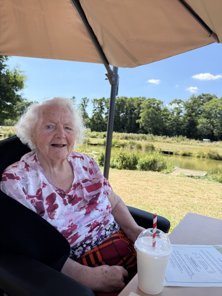 Elderly woman sitting outdoors under an umbrella holding a drink by a grassy riverside.