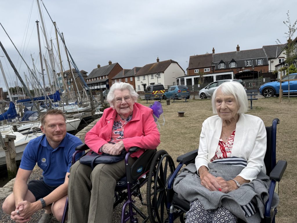 Two elderly women in wheelchairs with a caregiver sitting by a marina with boats.