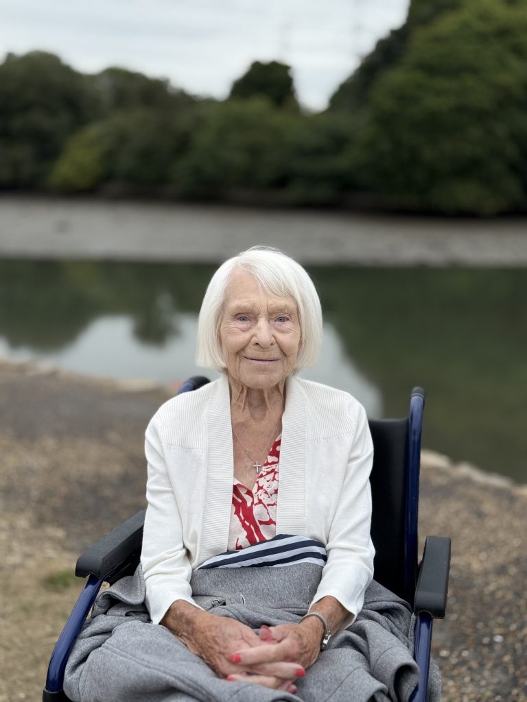 Elderly woman in a wheelchair smiling by a riverside with trees in the background.