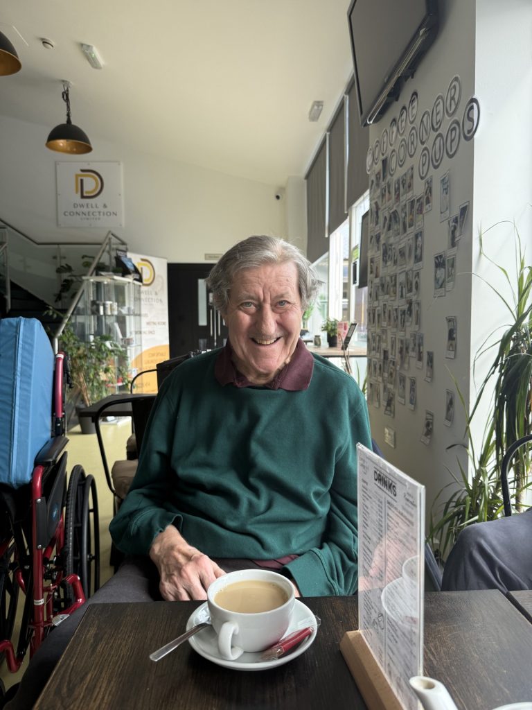 Elderly man smiling while having a cup of tea at a café table.