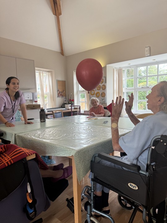 Caregiver and elderly residents playing with a red balloon around a table indoors.
