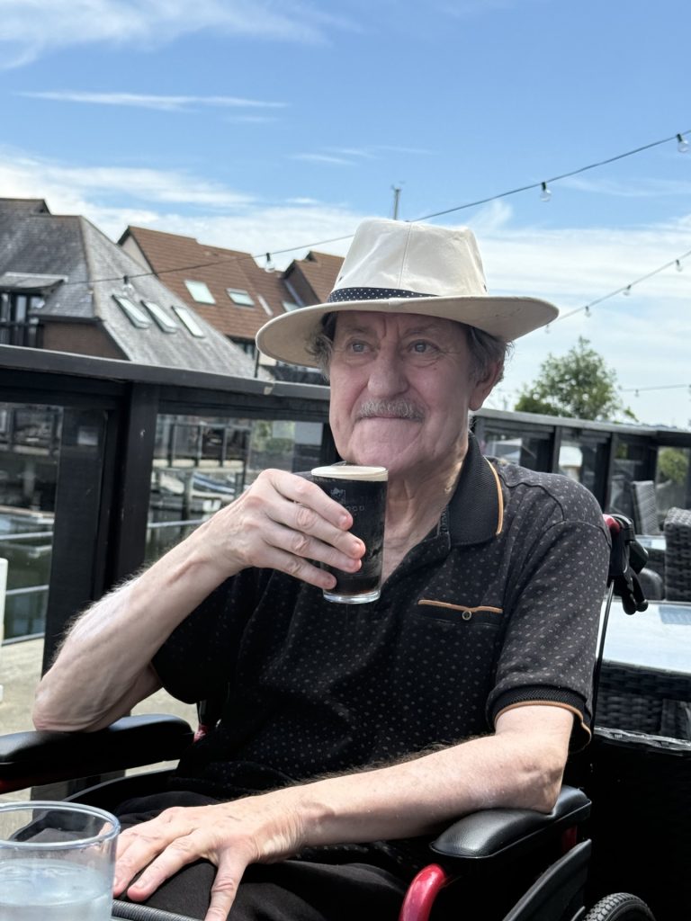 Elderly man in a hat sitting outdoors, holding a drink and smiling on a sunny day.