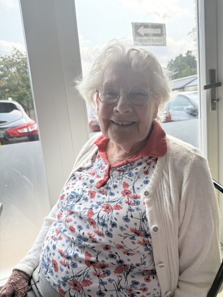 Elderly woman smiling while sitting by a window, wearing glasses and a floral top.