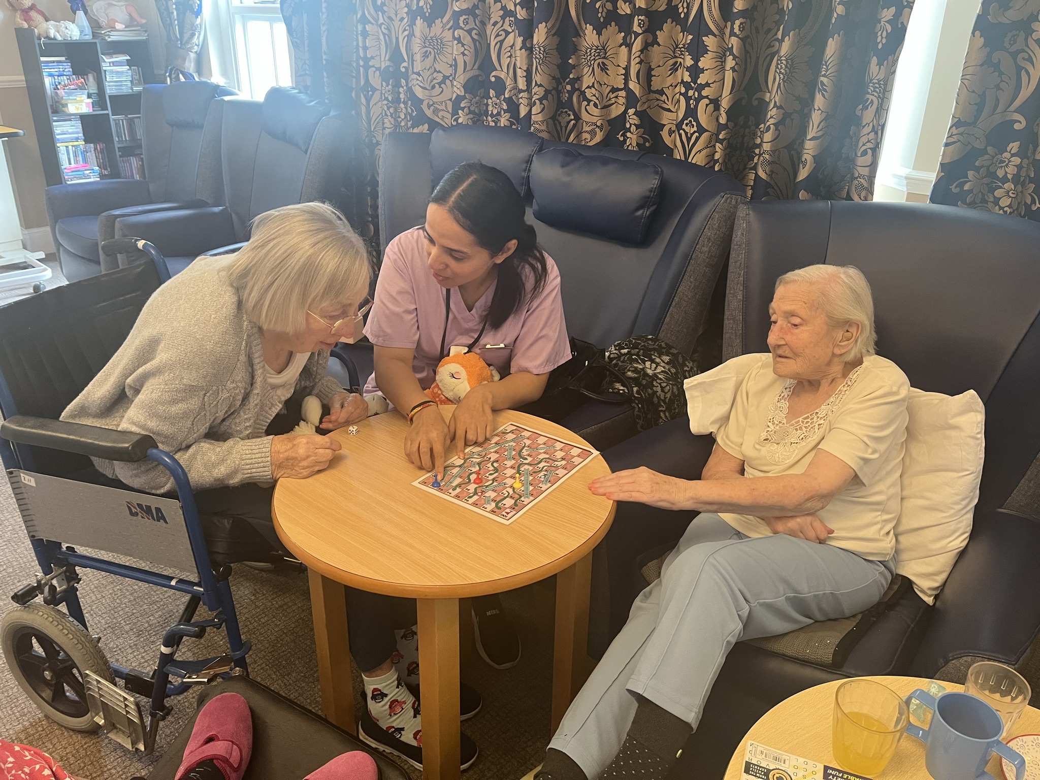 Two elderly women and a caregiver playing a board game together in a lounge.
