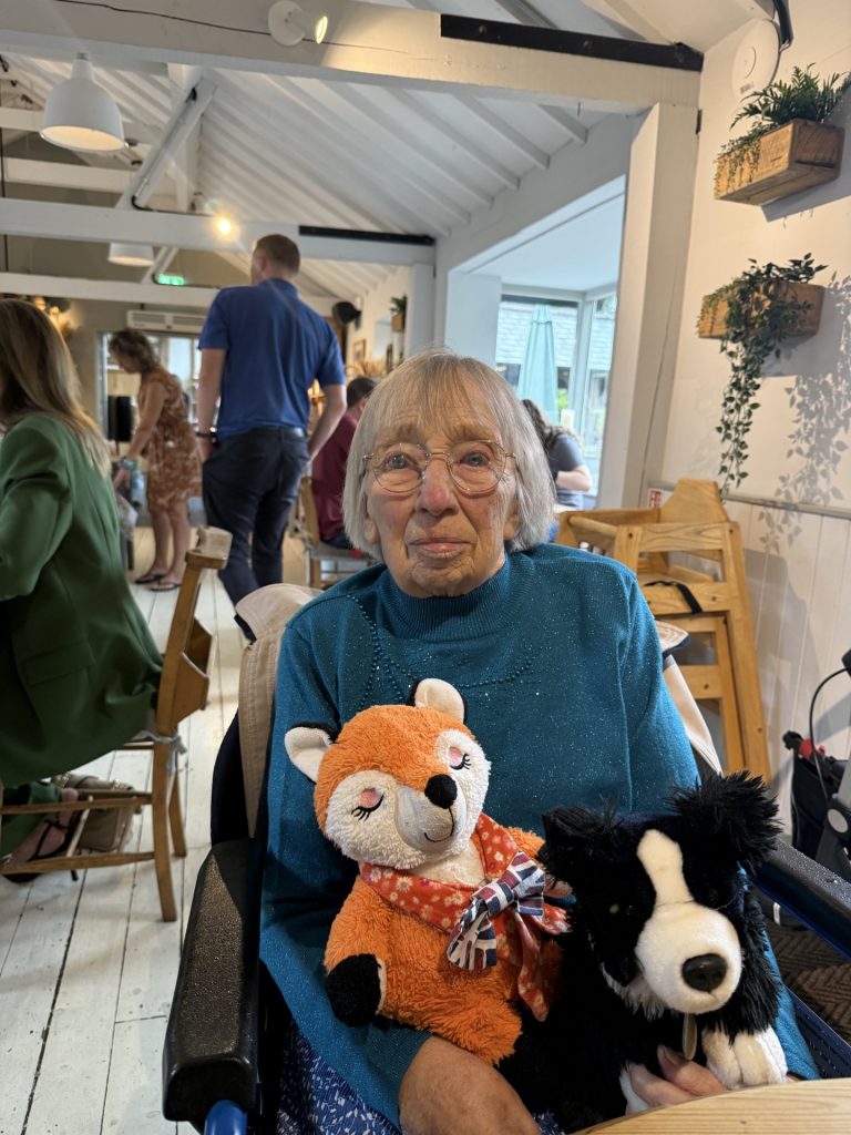 An elderly woman holding two stuffed animals, sitting indoors at a social gathering.