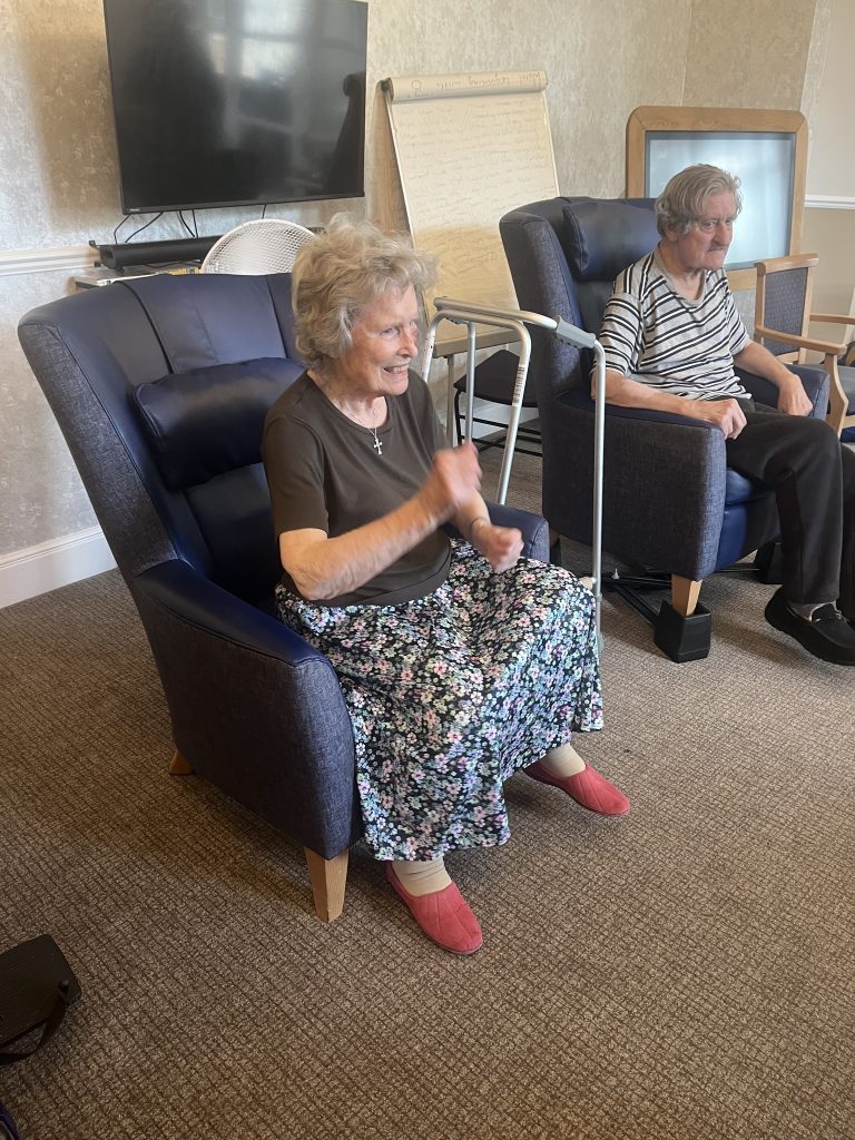 An elderly woman smiling and moving her hands while seated, with another resident sitting beside her.
