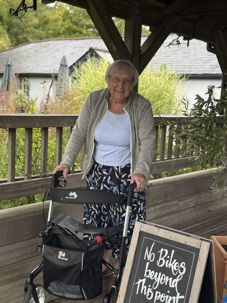 An elderly woman smiling while standing with a walker on a covered outdoor walkway.
