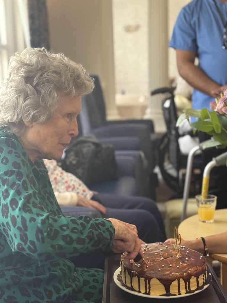 An elderly woman cutting a decorated cake while seated, with a drink and others nearby.