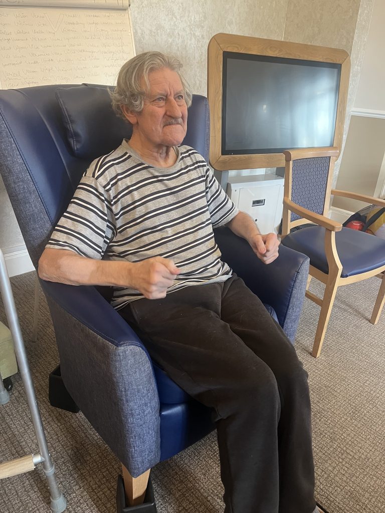 An elderly man seated in a chair, moving his arms as if exercising indoors.
