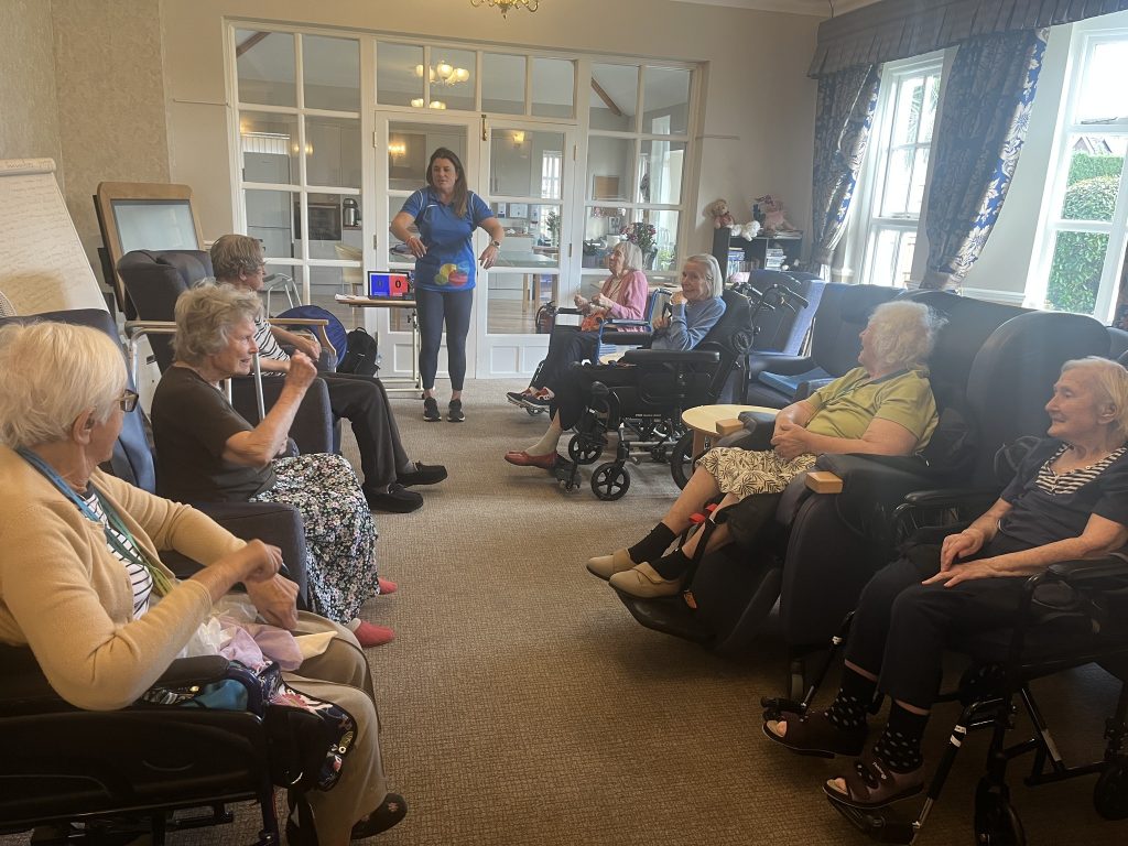 A group of elderly people seated in a circle participating in a guided activity with a caregiver in a lounge.