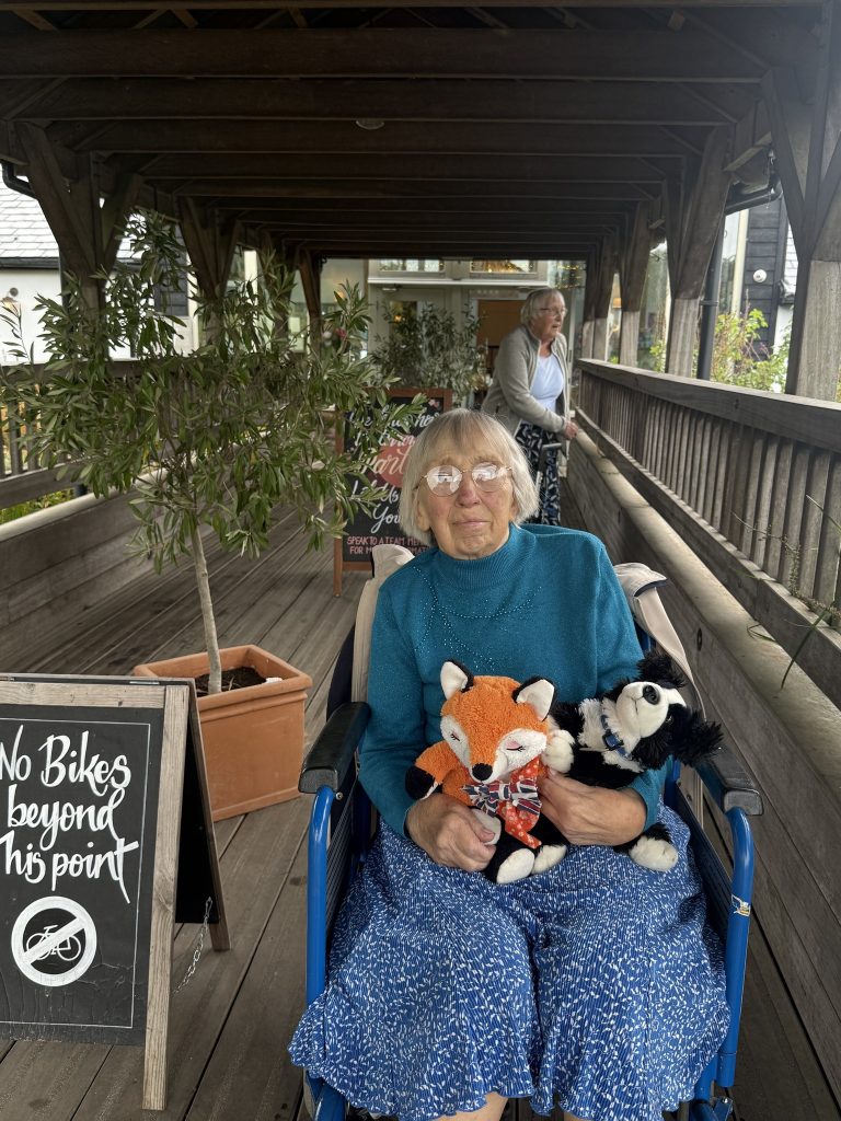An elderly woman in a wheelchair holding two stuffed animals on a wooden walkway, with another woman behind her.