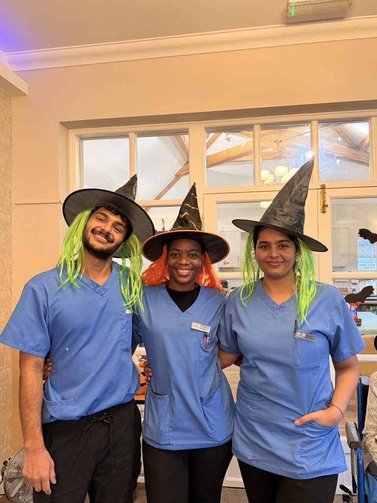 Three staff members in scrubs wearing witch hats and colorful wigs, smiling together.