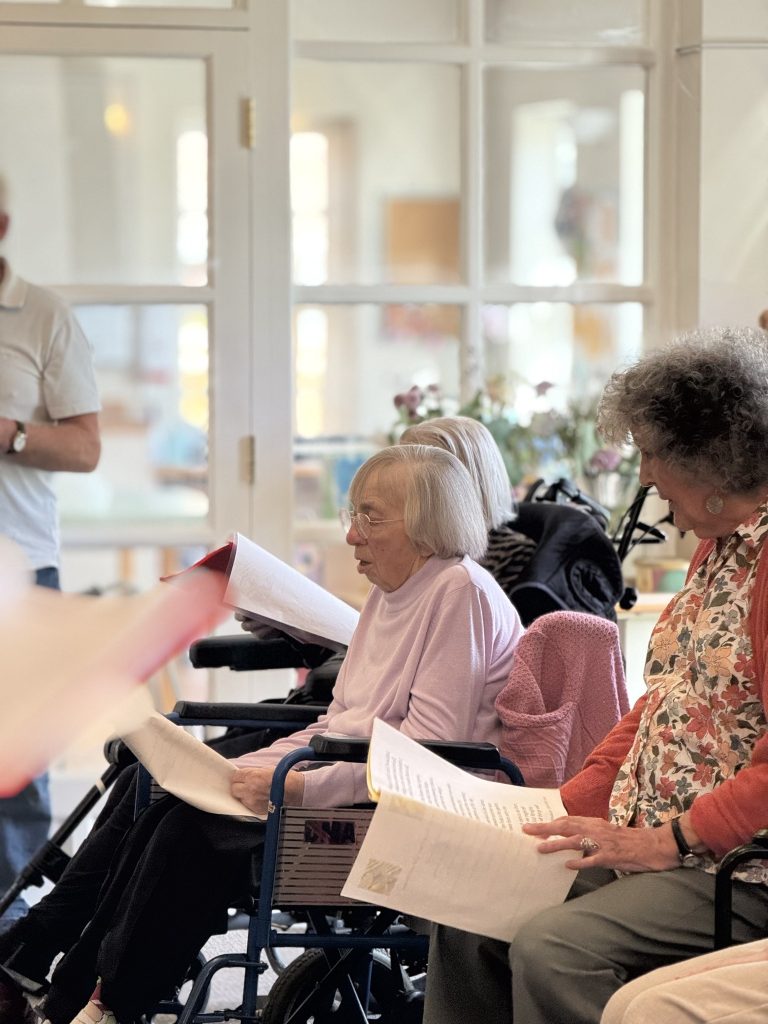 Group of elderly residents seated with papers, taking part in a reading or singing activity.