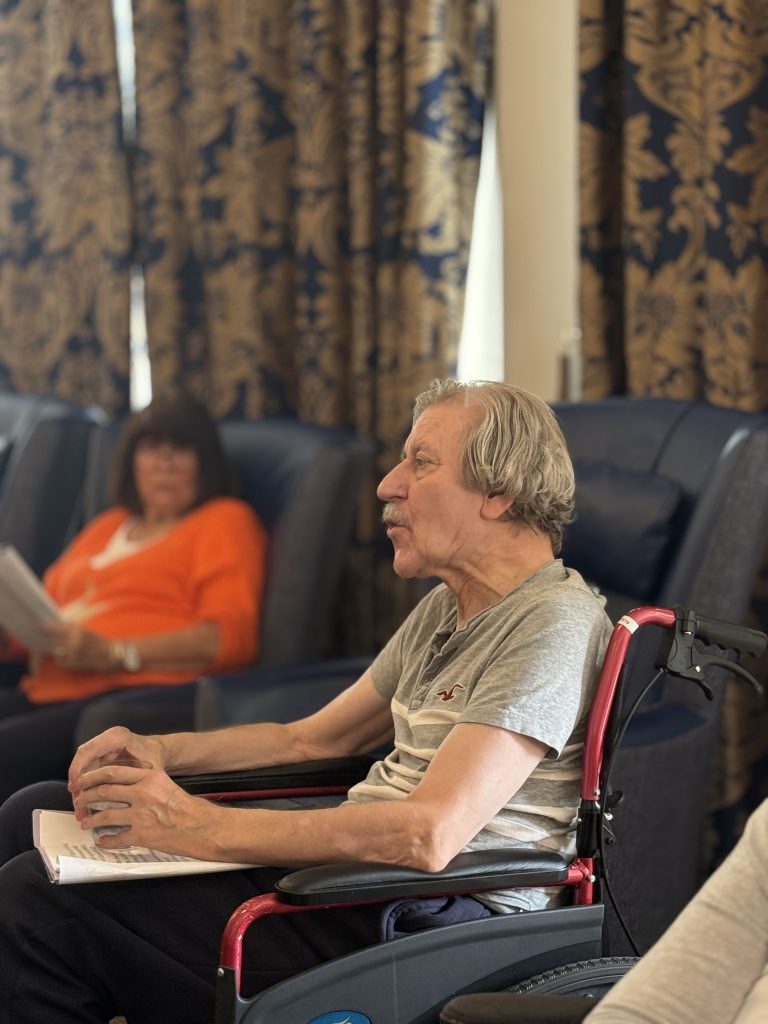 An elderly man in a wheelchair speaking during a group activity indoors.