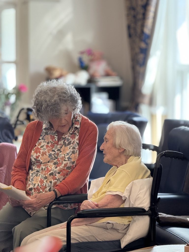 Two elderly women indoors, one showing a paper to the other as they talk.