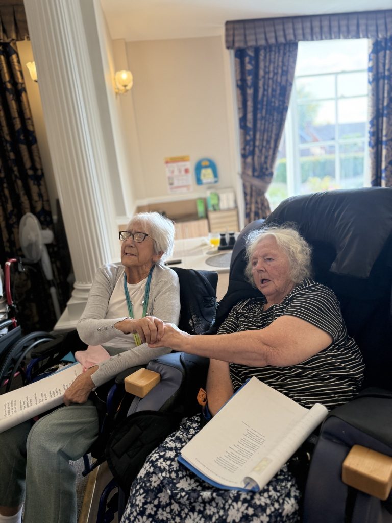 Two elderly women seated together holding hands, with papers in their laps, appearing engaged in an activity.