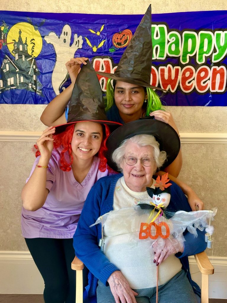 Two caregivers in witch hats posing with an elderly woman during a Halloween celebration.
