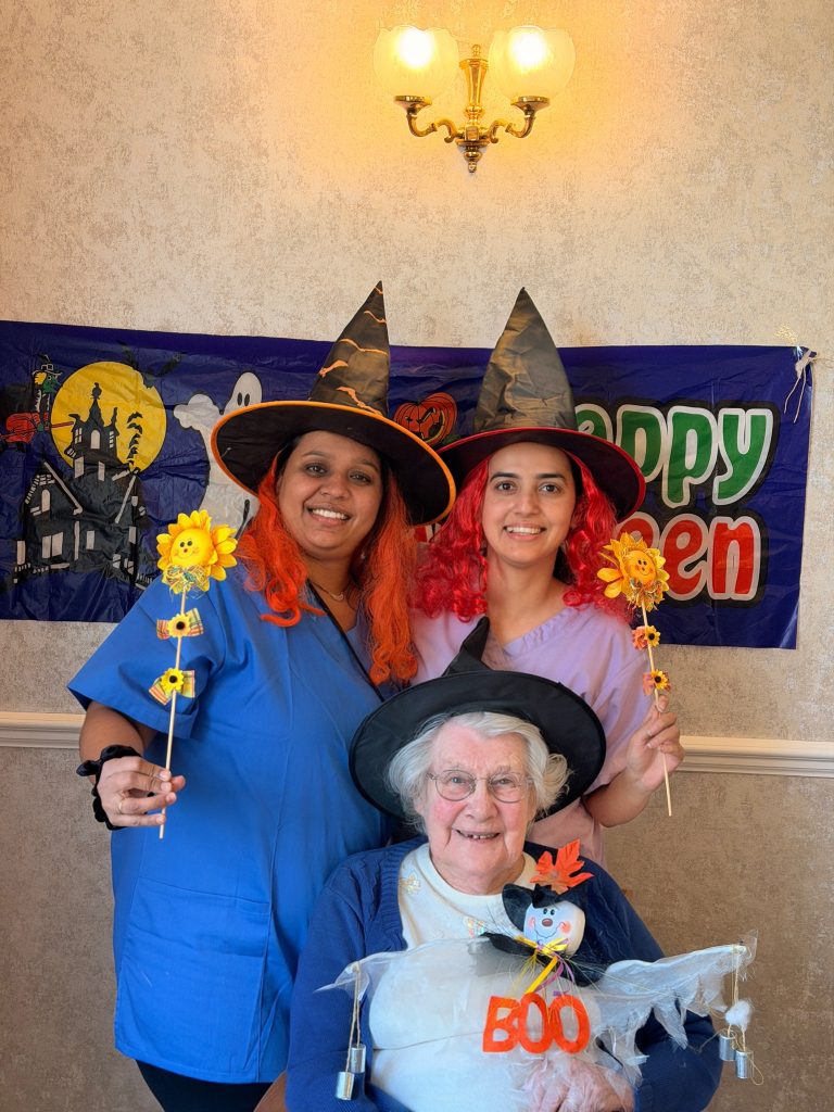 Two caregivers in costumes posing with an elderly woman under a Halloween banner.