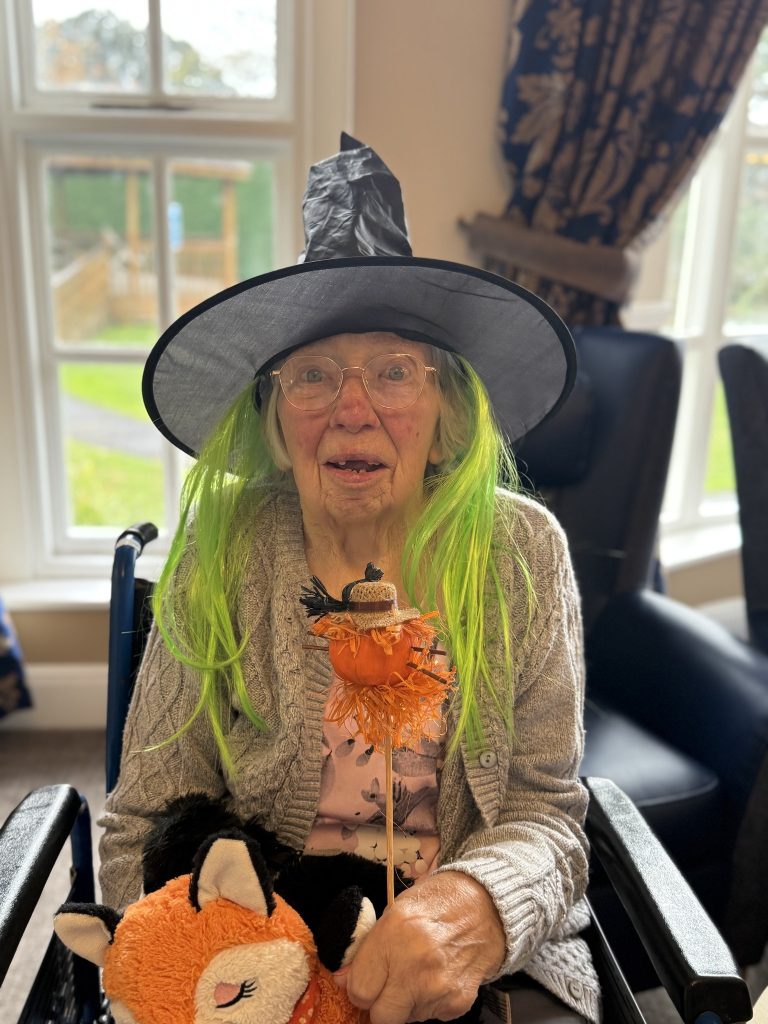 Elderly woman in a witch hat and green wig holding a Halloween decoration.