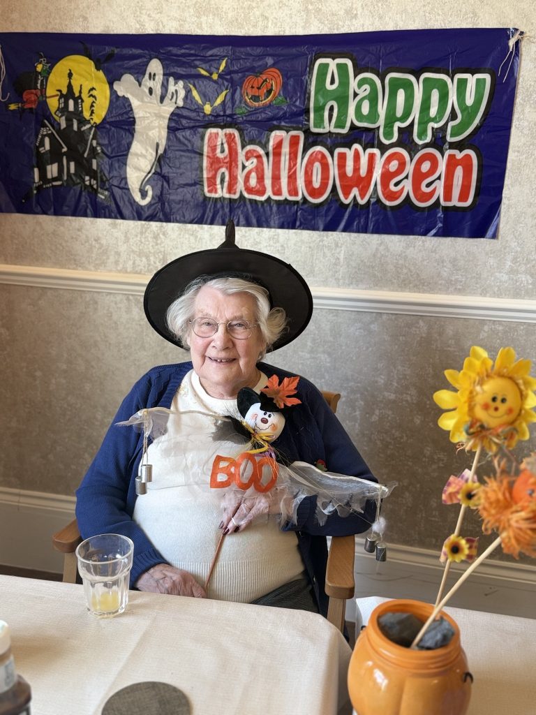 Elderly woman in a witch hat smiling under a “Happy Halloween” banner.
