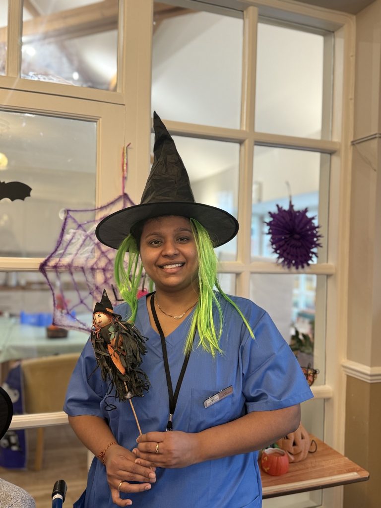 Care worker in scrubs wearing a green wig and witch hat holding a Halloween decoration.
