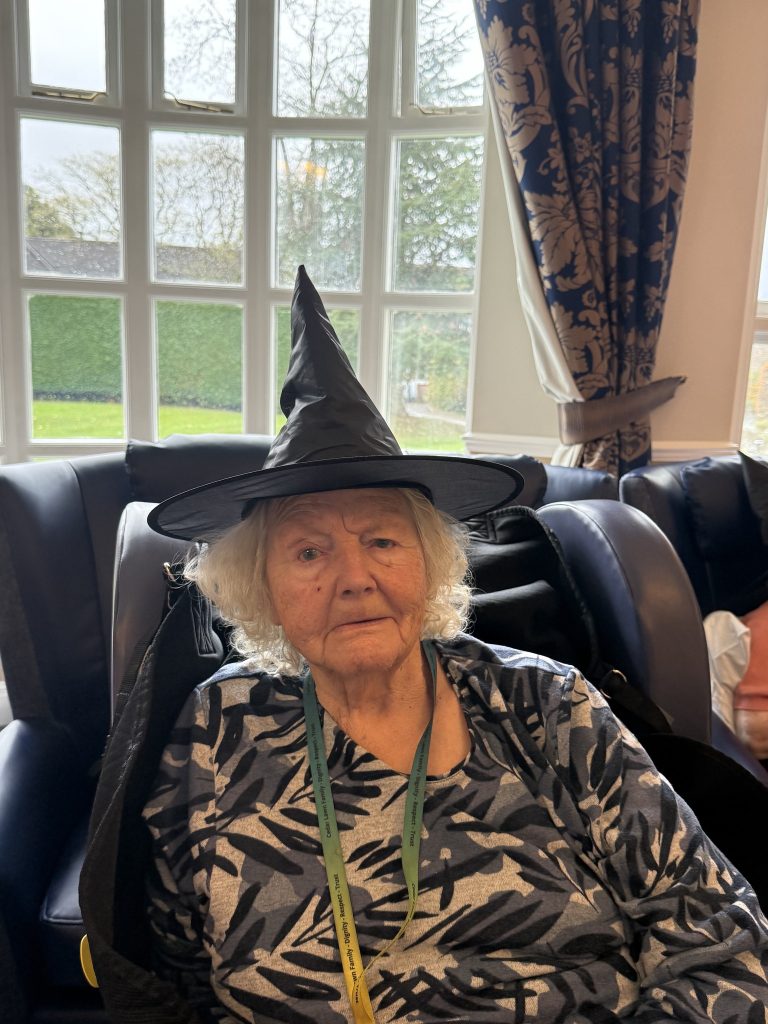 Elderly woman in a witch hat sitting by a window in a cozy room.