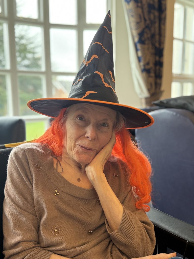 Elderly woman wearing a witch hat and bright orange wig, smiling with her hand on her cheek.
