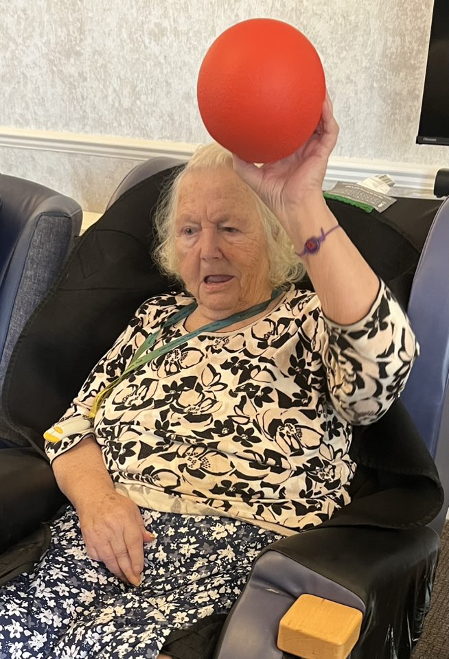 Elderly woman seated in a chair raising a red ball during an activity.