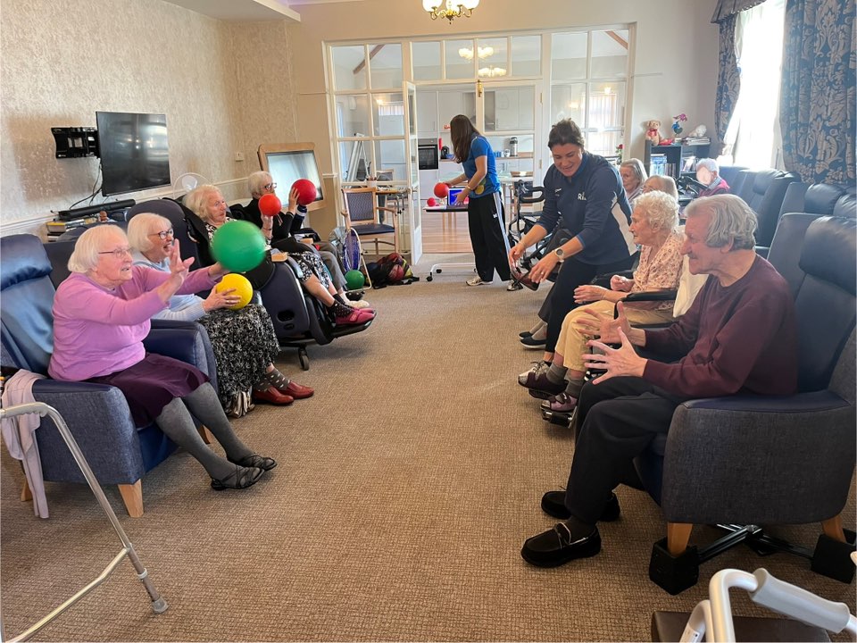 Group of elderly residents playing a ball game with staff in a communal lounge.