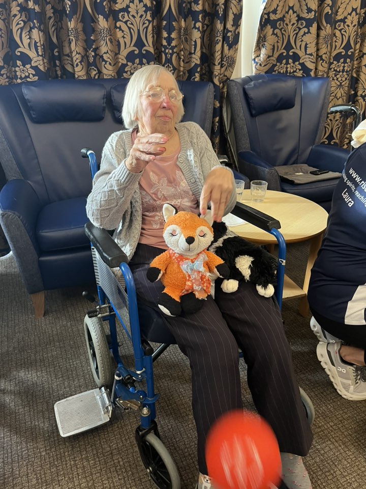 Elderly woman in a wheelchair with a toy on her lap, reaching toward a red ball mid-play.